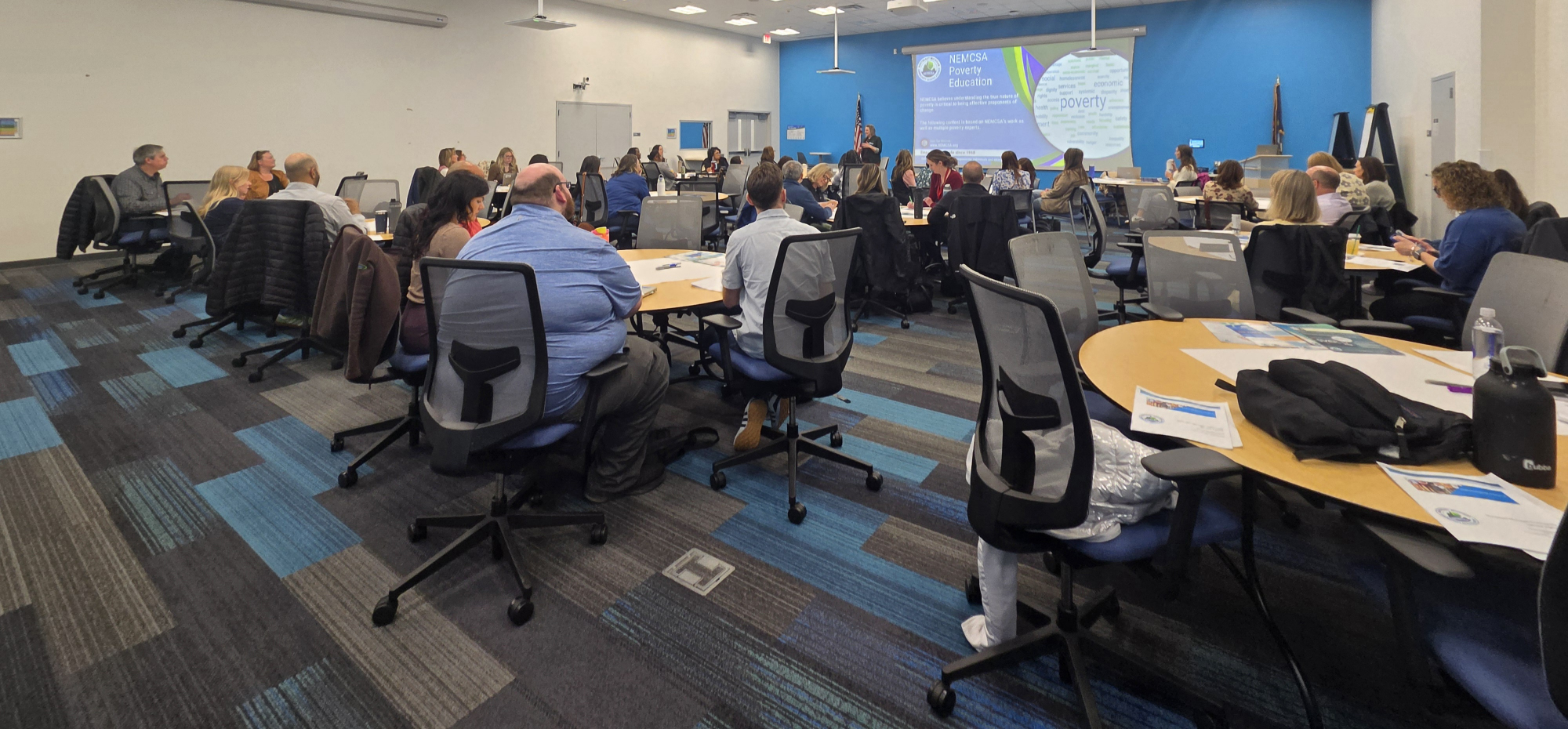 A large training room filled with Consumers Energy staff seated at round tables, engaged in NEMCSA's Poverty Education training in Jackson, MI. At the front of the room, a presenter stands near a projected "NEMCSA Poverty Education" slide on a blue accent wall. Training materials are visible on the tables. The room is full, reflecting strong attendance and engagement.
