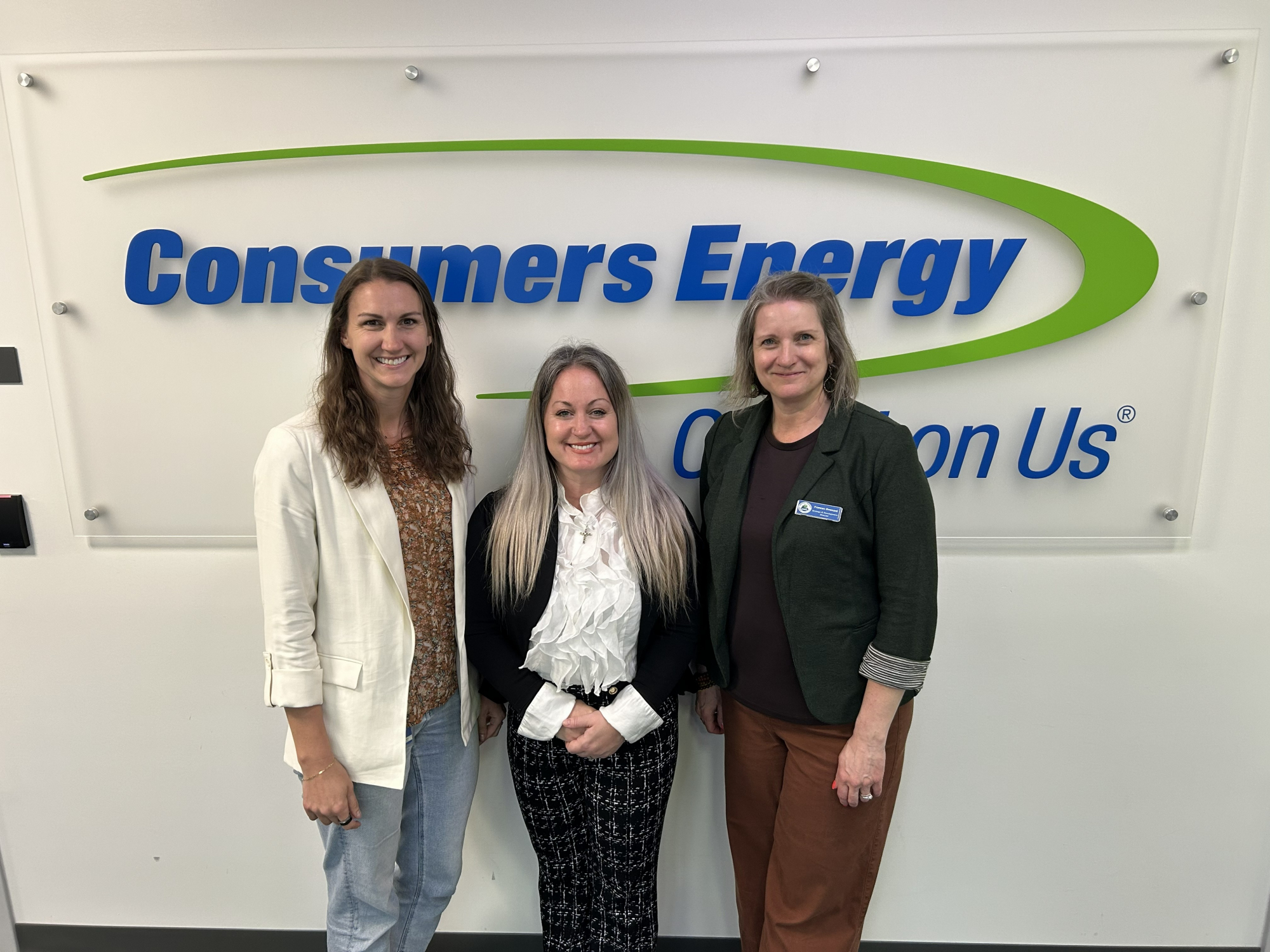 Three women stand smiling in front of a Consumers Energy lobby sign. From left to right: Frances Ommani (NEMCSA Strategy & Development Director), Kristen Goodman (Consumers Energy Manager of Energy Assistance Programs), and Amanda Bergeron (NEMCSA Community Resource & BOSS Program Manager), photographed at the Consumers Energy location in South Haven, MI.